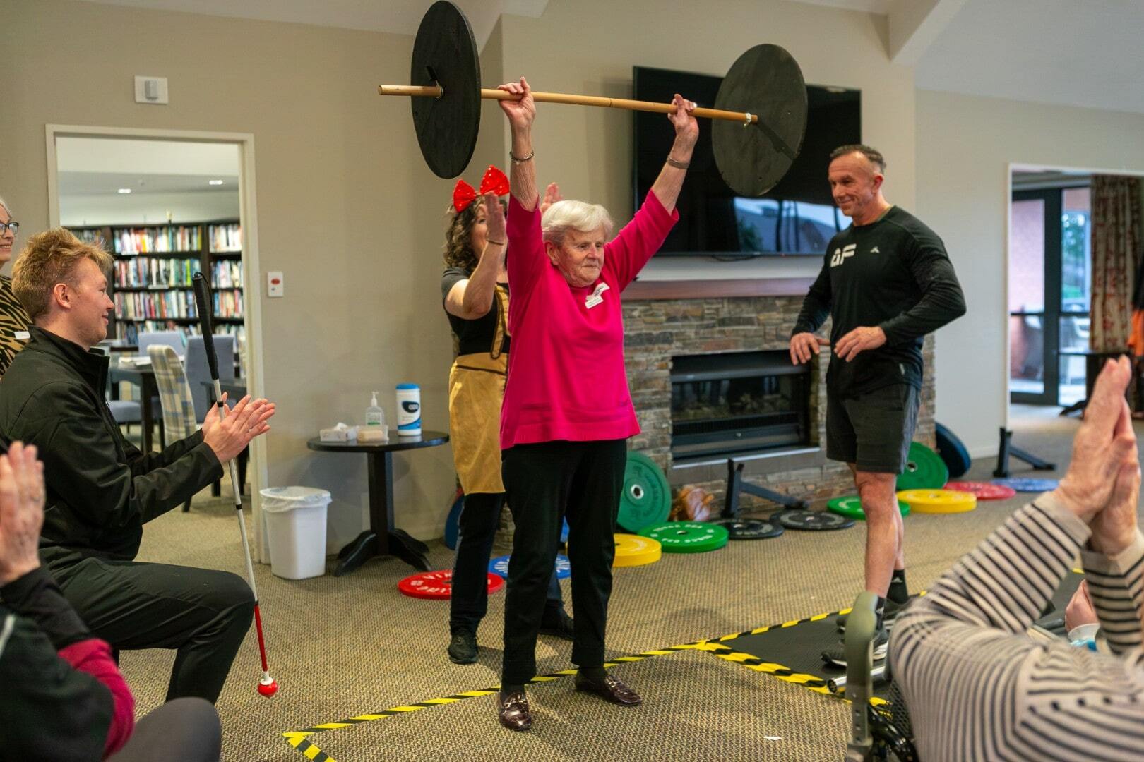 An older woman lifts a light barbell above her head as others look on enthusiastically