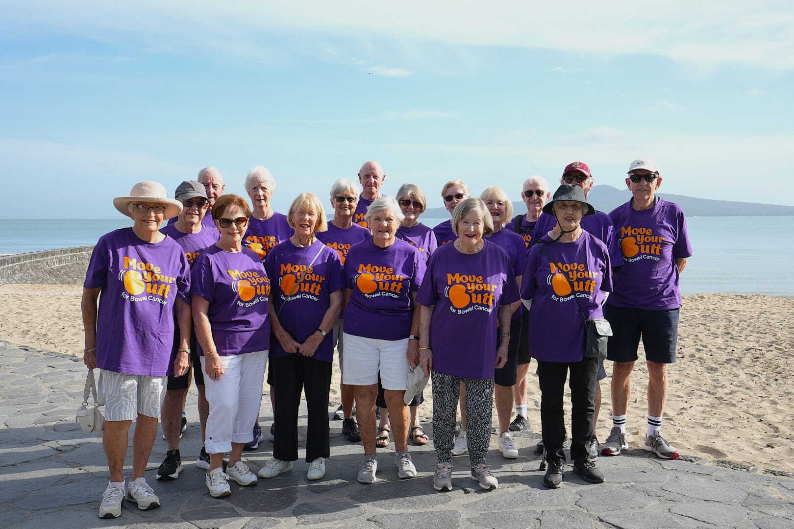 A group of older people stand by the beach wearing matching purple t-shirts