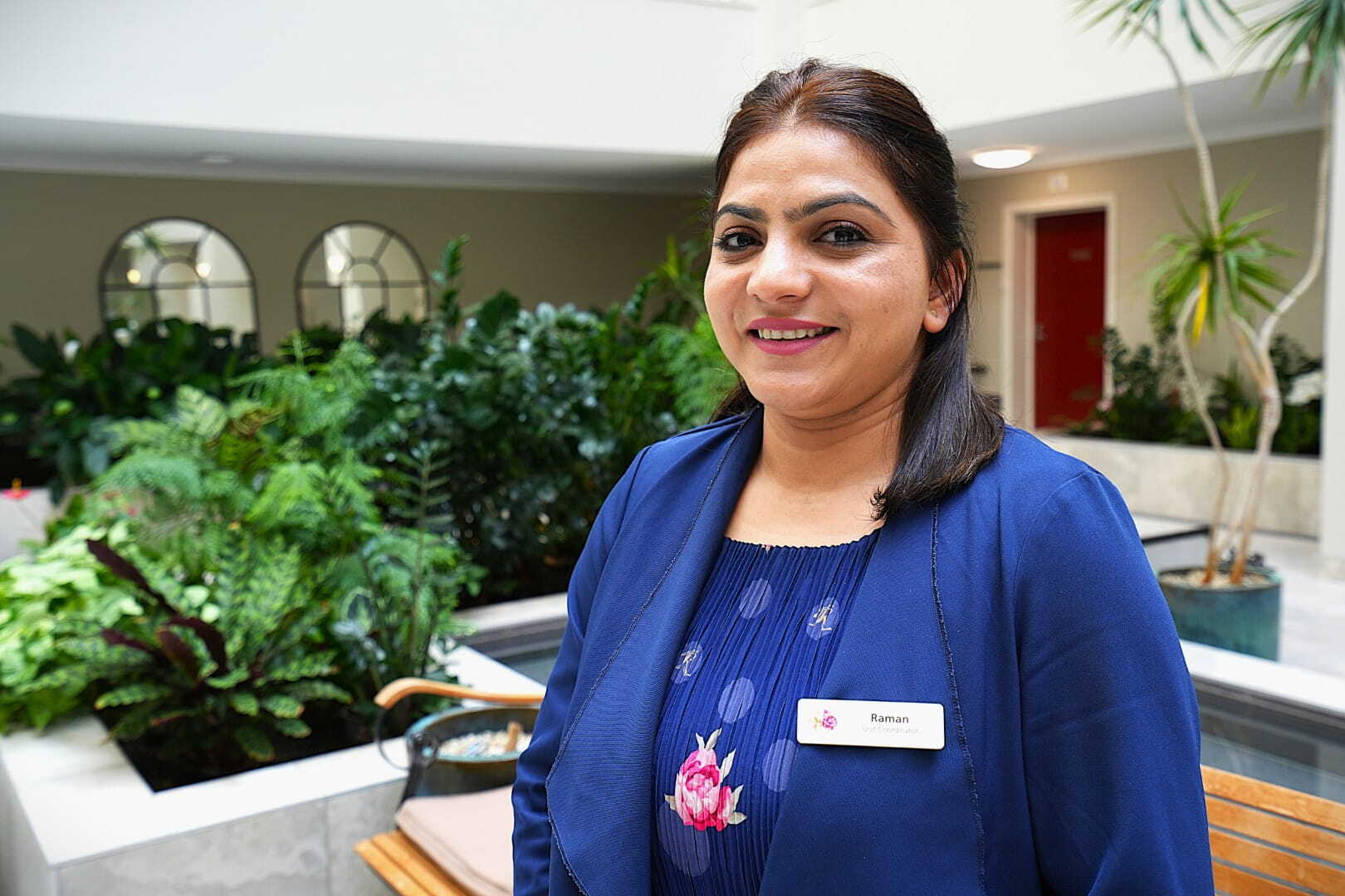 A smiling woman in uniform stands in a pleasant indoor environment with plants in the background