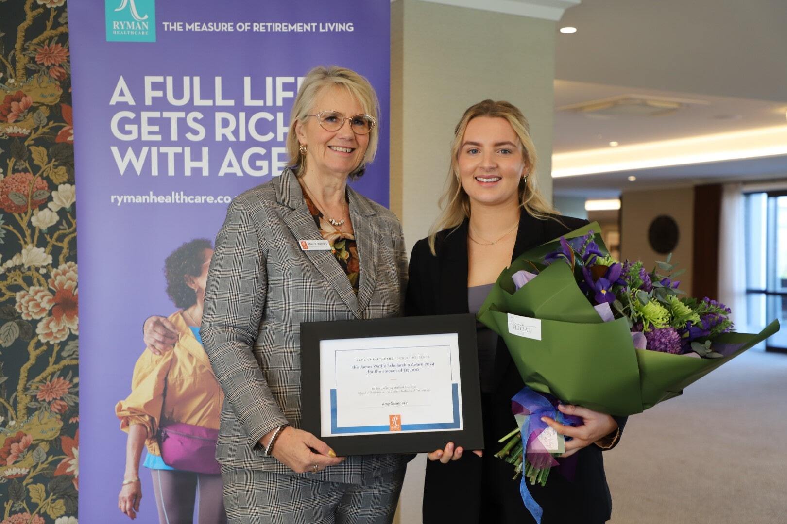 A young woman is awarded a framed certificate and a bouquet by an older woman