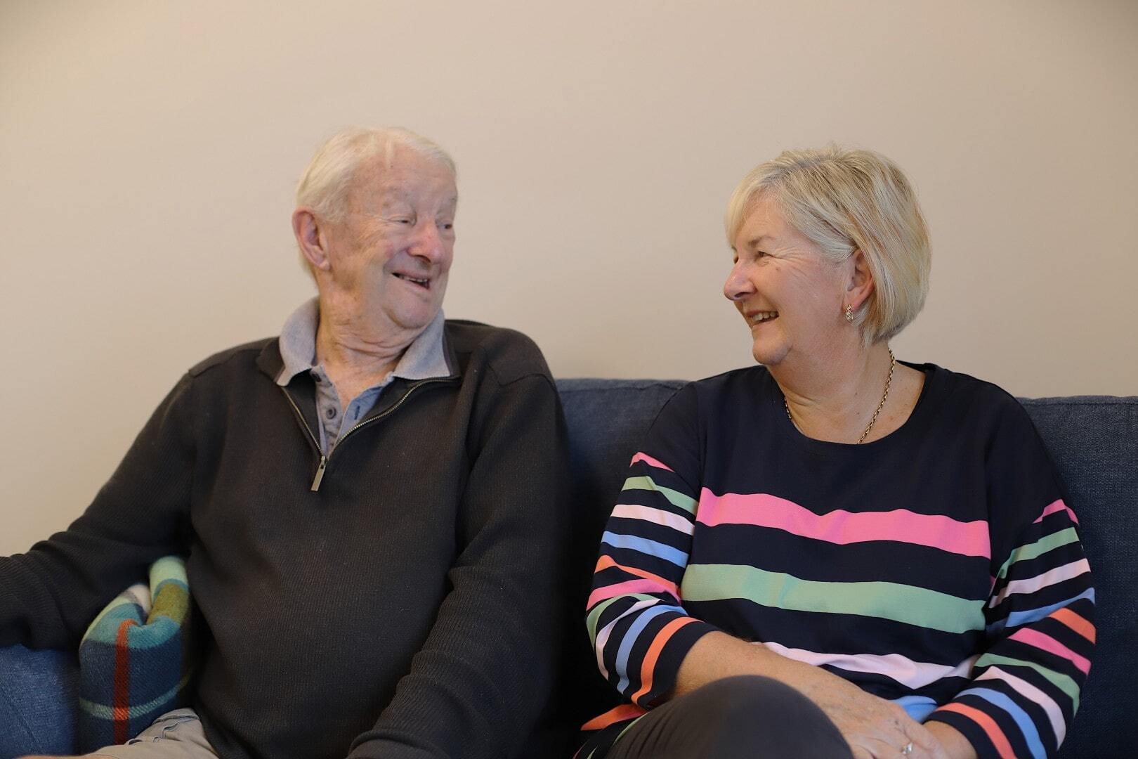 An older man sits on a couch with his middle-aged daughter, smiling.