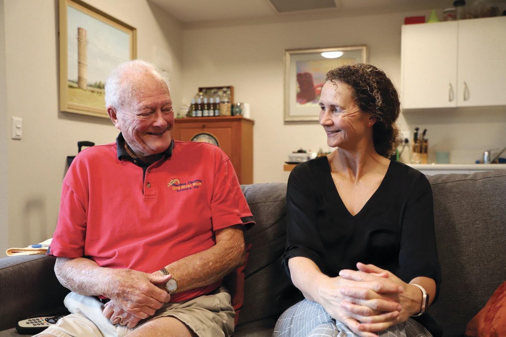Older man sits on a sofa next to his middle-aged daughter, both smiling