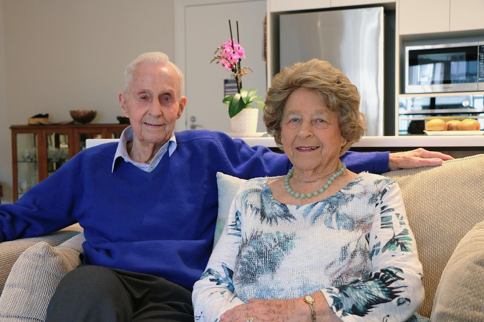An older couple sit together on their couch smiling at the camera