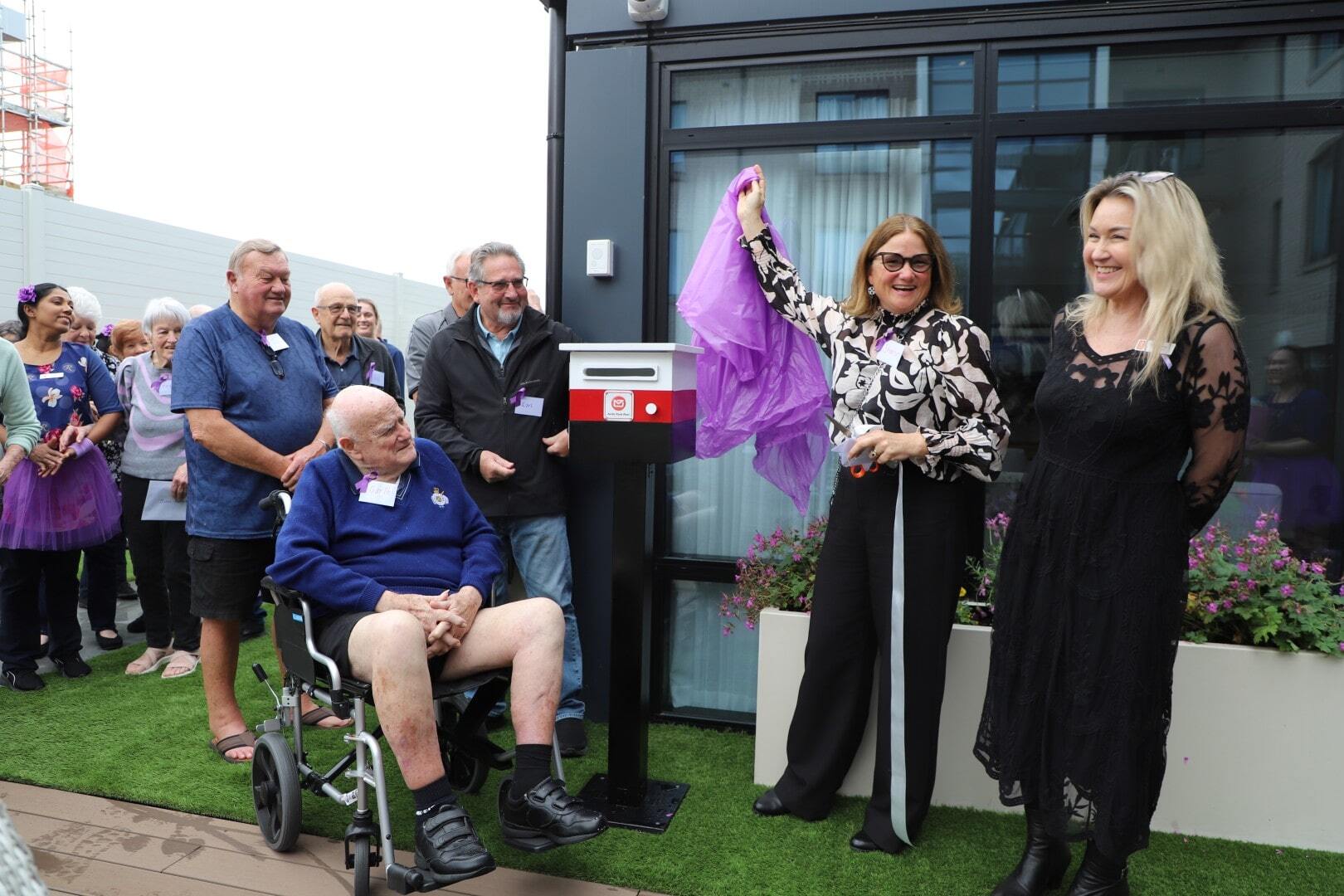 A smiling woman unveils the cover off a mailbox while others watch on in delight