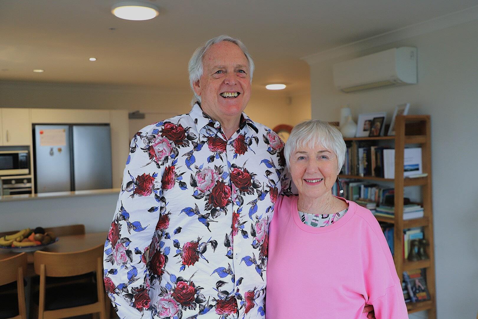 An older couple dressed in patterned and bright coloured clothing smile in their stylish apartment