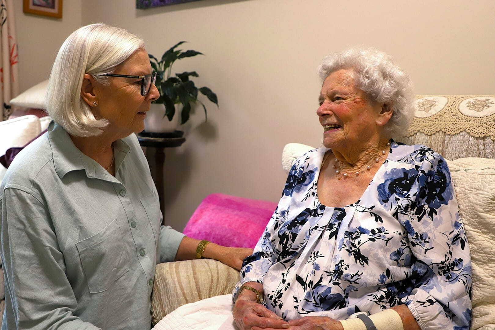 An older woman sits in a chair and smiles at her daughter in a cosy room