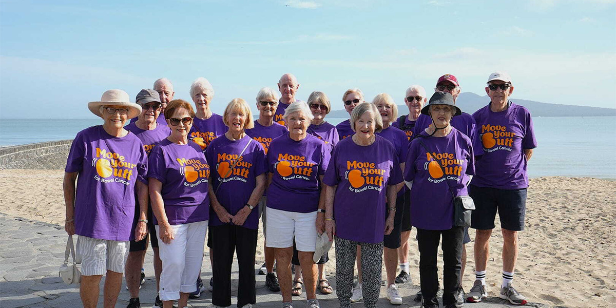 A group of older people stand by the beach wearing matching purple t-shirts