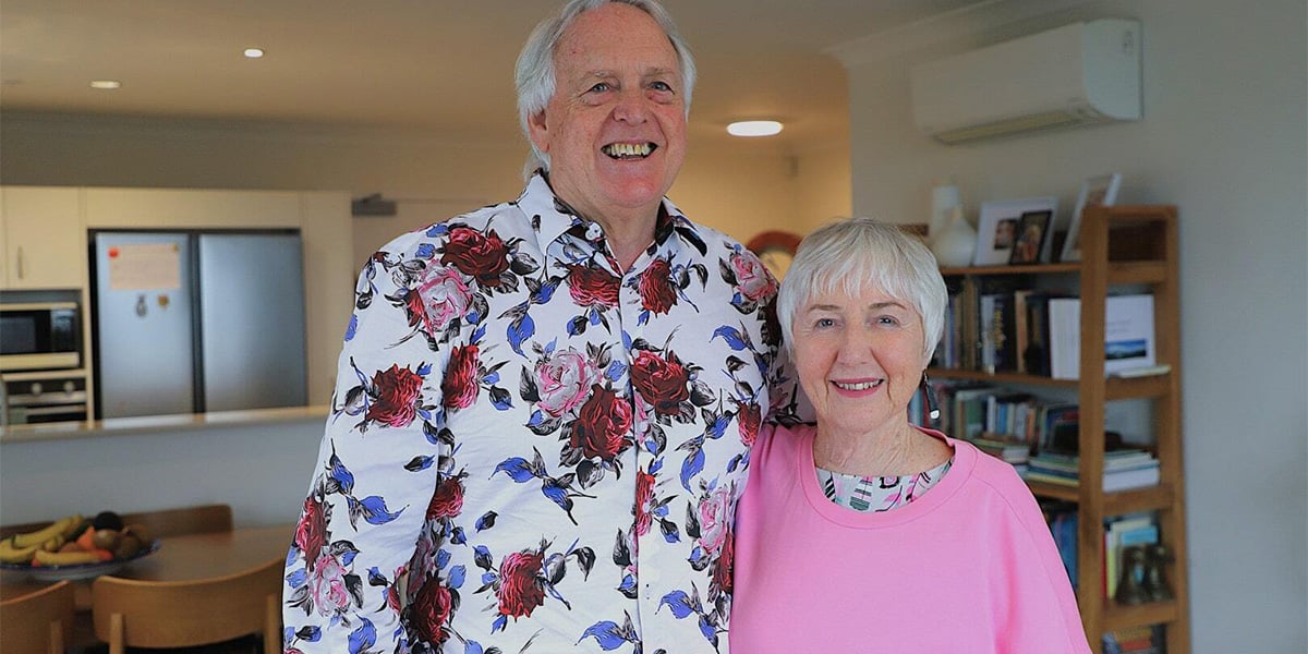 An older couple dressed in patterned and bright coloured clothing smile in their stylish apartment